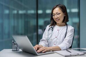 Asian doctor working on a laptop in her office. She is wearing a white coat and stethoscope while smiling and typing on her computer photo