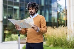 Man wearing brown sweater reads map outside office building. Focused expression suggests navigation or planning. Surrounded by greenery, holding suitcase, indicative of travel or business trip. photo