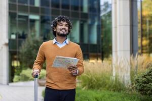 Young businessman holding map and suitcase outside modern office building. He looks ready to embark on travel journey, exuding confidence and curiosity. Perfect portrayal of urban exploration. photo