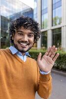 Smiling businessman in casual attire waving at camera during call using phone. Image captures friendly, cheerful mood in urban business setting, with modern office in background. photo