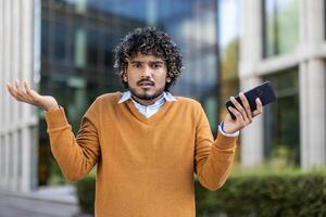 Man with curly hair in sweater holding smartphone with confused expression. Outdoor urban background suggests business context, conveying uncertainty, doubt, or perplexity in modern communication. photo