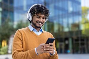 Smiling young man using smartphone with headphones in urban environment. Image captures casual lifestyle, technology interaction, and modern communication. photo