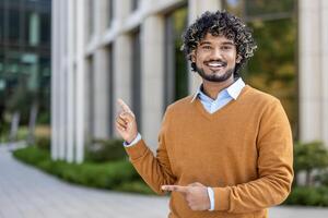 Smiling young professional stands outside modern office building, confidently pointing with both hands. Image conveys positivity, success, and enthusiasm in business or academic environment. photo
