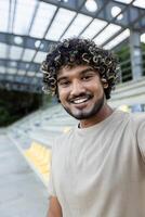 A young man smiles into the camera while calling at the stadium, in a casual setting with seats visible in the background photo