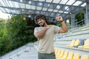 The athlete was injured while engaging in active physical exercises and fitness. The man is massaging the shoulder and back muscles, standing outside in a park photo