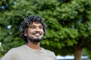 A young man with curly hair and a short beard is smiling and looking up against the bokeh background of lush green trees. photo