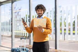 Tourist at public transport stop with phone in hands and map, smiling man uses app while standing with suitcase. photo