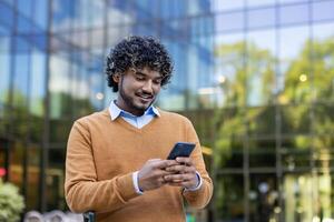 Confident young man engaging with smartphone outside office building. Wearing casual attire, he appears focused and content. Represents modern communication, professional networking. photo