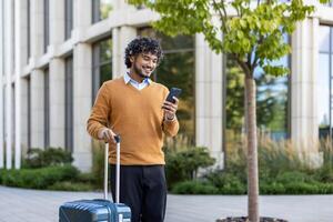 Smiling man checks phone while pulling suitcase outside modern building. Represents business travel, technology use, and professional lifestyle in casual attire, connecting work and leisure photo