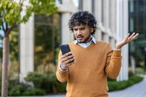 Young man appears perplexed as he looks at smartphone, expressing confusion and frustration. Dressed in casual attire with orange sweater, he stands outside in urban environment photo