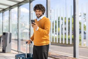 A smiling man looks at his phone while waiting at a bus stop, luggage in hand. He is dressed in a sweater and a collared shirt photo