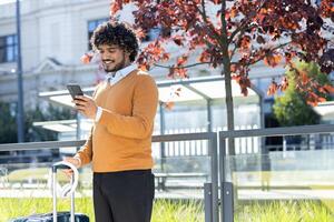 Young man waiting for public transport standing at a stop with a suitcase. Tourist smiling happily using an app on his smartphone. photo