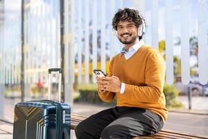 A smiling man with headphones uses his phone while waiting at a bus stop, a suitcase beside him. He is dressed casually and looks relaxed photo