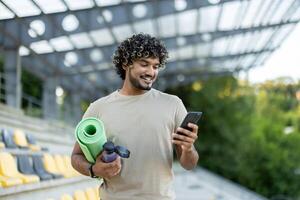 Male athlete with yoga mat and phone walking in the park. Using smartphone app for training and viewing physical exercises. photo