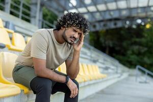 Tired and overworked athlete sitting on a bench, man resting after active physical exercises, fitness and jogging. Suffering from headache. photo