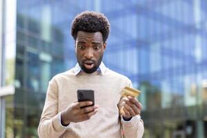 Deceived man standing outside office building with phone in hand and bank credit card. Businessman received rejection and error of online money transfer. photo