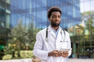Portrait of a young confident and serious doctor. A man with a tablet computer in his hands and in a white medical coat looks focused at the camera while walking outside the clinic office. photo