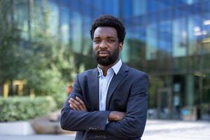 A confident Black businessman stands with crossed arms in front of a modern office building with blurred greenery. photo