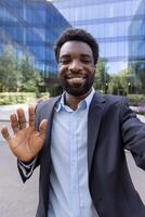 A young, handsome man in a blazer smiles and waves to the camera during a mobile call in front of a modern office building photo
