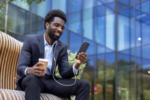 Young businessman is enjoying a coffee break while talking on his cell phone sitting on a modern bench near the office building. photo