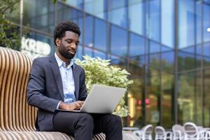A young professional works remotely, utilizing his laptop while seated on a modern bench with an office building as a backdrop. photo