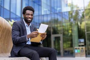 A smiling businessman opens an envelope, possibly containing good news, while sitting outside an office building on a modern bench. photo
