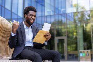 A young businessman cheers with his fist raised while sitting outside, having just opened a letter with great news in front of an office building photo