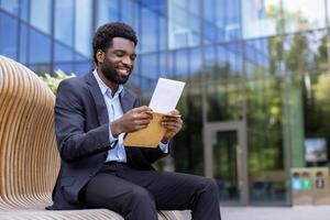 An african american businessman in a suit sits outside, smiling while reading a document in an envelope, happy. The setting is a modern office environment. photo