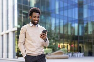 An african-american man stands in front of a glass building, looking at his phone with a pleasant expression. It's a sunny day. photo