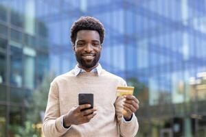 A cheerful man holding a smartphone and credit card, smiling towards the camera in an outdoor setting. He is likely engaged in a transaction. photo