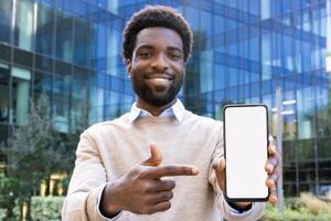 A smiling Black man in front of a modern building points to a blank white screen on his mobile phone, suggesting technology. photo