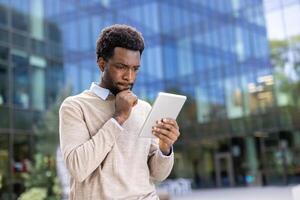A thoughtful businessman examines his tablet, appearing to ponder information with a hand on his chin, outdoors near a modern building. photo