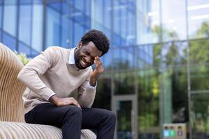 A black man is experiencing a painful moment while sitting on a modern bench outside a building. The man is clearly distressed. photo