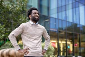 A smiling man with hands on hips poses confidently outside a modern glass building. The soft sunlight enhances the happy expression on his face. photo
