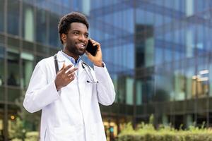 A smiling african-american doctor in a white coat talks on his phone outside a modern building. He gestures with his hand while conversing. photo