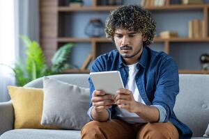 Young man with curly hair looks puzzled while browsing on his tablet in a modern living room setting. He appears surprised by something he sees. photo