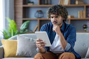 Serious thinking man using tablet computer reading e-book while sitting on sofa in living room at home. photo