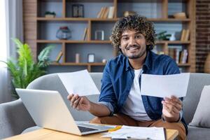 A smiling man is looking at the camera holding papers in front of a laptop while working remotely from home. photo