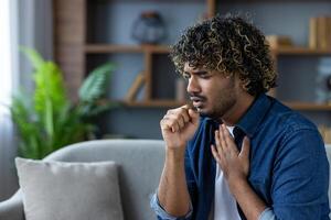 Man with curly hair coughing while sitting on a sofa in an interior setting, possibly experiencing symptoms of illness or allergy. photo