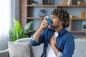 A young man with curly hair is using an inhaler for his asthma while sitting on a sofa in a bright living room. photo