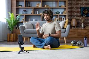 A smiling man waves while conferencing at home during his fitness routine. The scene includes a yoga mat and dumbbells. photo