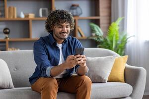 Young successful man using app on phone while sitting inside apartment on sofa at home. Satisfied smiling typing text message and browsing social media. photo