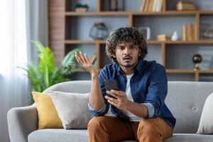 A young man looks confused while holding his smartphone, questioning a message or notification, seated on a couch in a home setting. photo