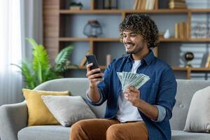 Happy man checking bank account balance using a mobile app and holding a fan of cash, sitting on the couch at home. photo