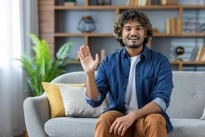 A cheerful young man with curly hair is waving with a smile while sitting on a sofa in his living room, looking into the camera. photo