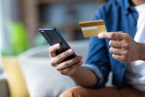 Man is holding a gold credit card and smartphone while sitting indoors. He is about to make an online payment using his mobile device. photo