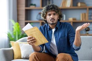 A confused man sits on a sofa at home, shrugging while holding a letter he just opened, with a bookshelf visible in the background. photo
