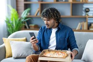 A man looks at his phone with a confused expression while holding a pizza box, possibly dealing with an order issue photo
