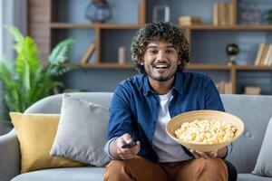 A happy man with curly hair watches television while relaxing on the sofa with a large bowl of popcorn at home. photo