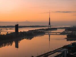 Serene sunrise over Riga, Latvia, featuring the Riga TV Tower and its reflection in the Daugava River, with a bridge and modern high rise visible. photo